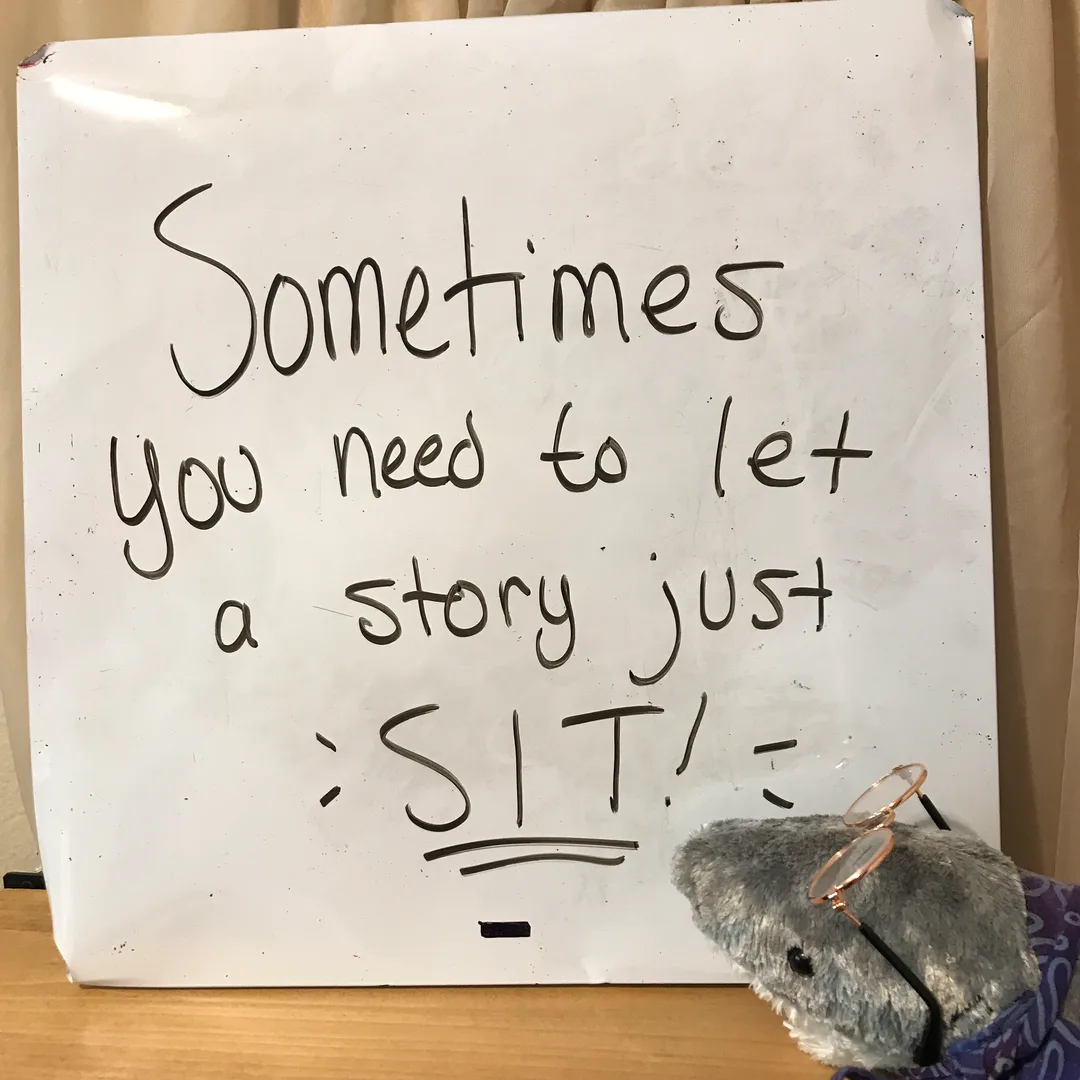 A photograph of a plush shark with a scarf and glasses sitting beside a white board that says 